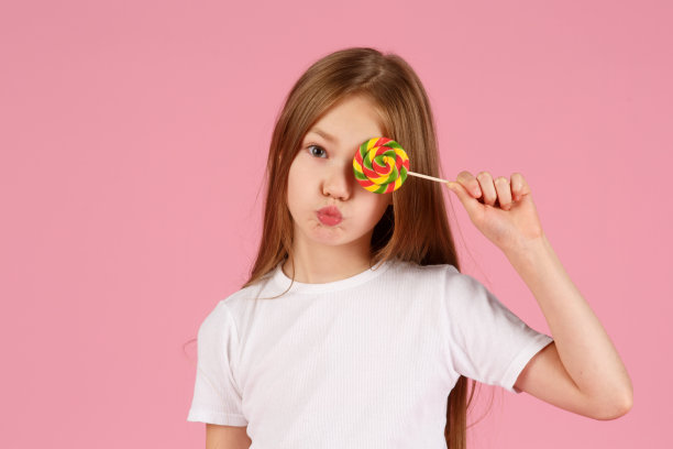 一个惊讶的小女孩的肖像拿着心形棒棒糖，看着孤立的相机在粉红色的背景图片下载