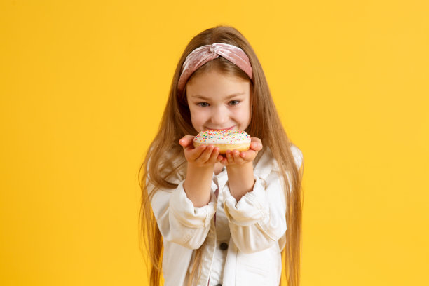 疯狂快乐的金发女孩戴着生日帽微笑，玩得很开心，看着她眼睛上的两个红色甜甜圈。糖果。黄色工作室背景图片下载