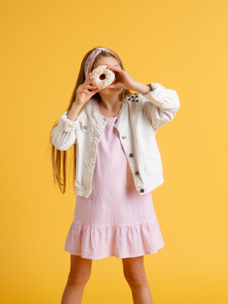 疯狂快乐的金发女孩戴着生日帽微笑，玩得很开心，看着她眼睛上的两个红色甜甜圈。糖果。黄色工作室背景图片下载