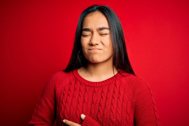 年轻美丽的亚洲女人穿着休闲毛衣站在孤立的红色背景与手对胃，因为消化不良，痛苦的疾病感觉不舒服。疼痛的概念。图片下载