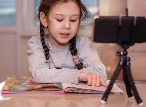 远程学习在线教育。女学生正在看书。用三脚架上的手机录制视频图片下载