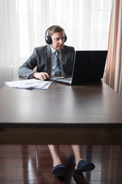 年轻的失业商人穿着灰色西装、衬衫和拖鞋在远程办公，因为冠状病毒大流行COVID-19的检疫。远程工作在家的桌子。通过笔记本电脑与老板进行耳机在线会议图片下载