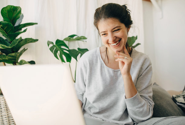 快乐的女孩在笔记本电脑上聊天，坐在有枕头和植物的现代房间里。家庭办公室。自由和自由。年轻随意的女人用笔记本电脑交流，观看网络研讨会图片下载