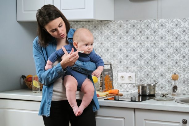 年轻的母亲抱着婴儿，用勺子喂蹒跚学步的儿子素材图片