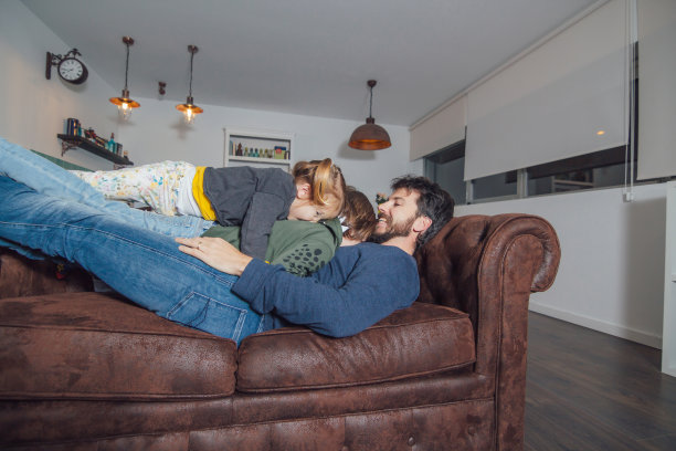 快乐的年轻父亲躺在沙发上，他的两个小孩在上面图片下载