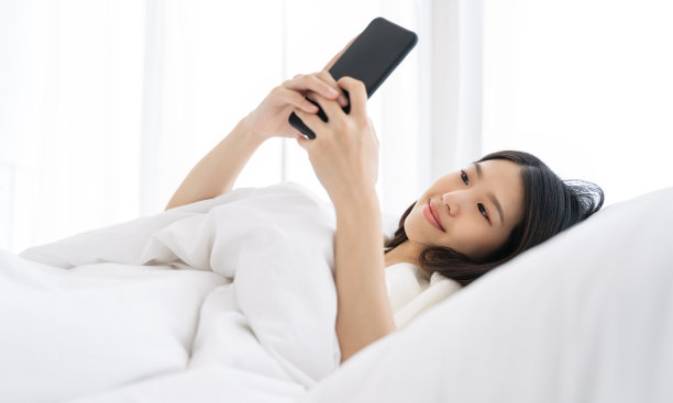 年轻的亚洲女人使用智能手机在卧室在家里快乐和微笑。图片下载