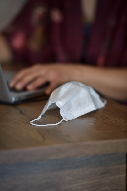 在家里工作的概念照片，在冠状病毒大流行时期，描绘了一个妇女在她的笔记本电脑上打字，旁边是一个保护性的医疗口罩图片下载