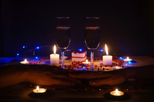 一对手的特写，桌子上有两杯香槟，蜡烛，心形，珠子。一对浪漫的情侣在晚餐时牵着对方的手。接触的概念。图片下载