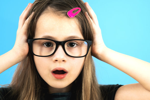 惊讶的小女孩的肖像戴着眼镜，手拿着她的脸孤立在蓝色的背景。图片下载