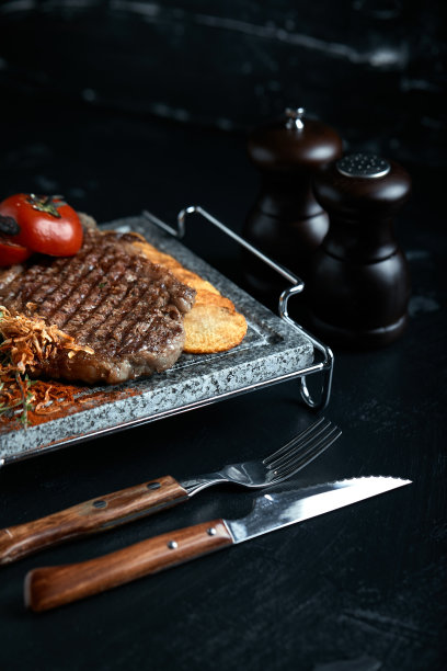 用刀叉雕刻在黑色石板上的烤牛排。在滚烫的大理石上烤牛排。复制空间，深色背景，美食时尚照片图片下载