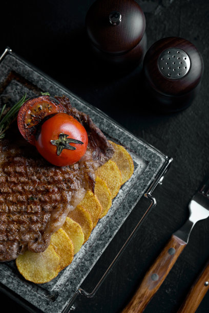 用刀叉雕刻在黑色石板上的烤牛排。在滚烫的大理石上烤牛排。复制空间，深色背景，美食时尚照片图片下载