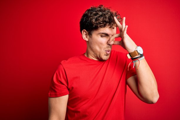年轻的金发帅哥卷头发穿着休闲t恤在红色的背景闻着一些恶臭和恶心，难以忍受的气味，屏住呼吸用手指在鼻子上。不好的气味图片下载