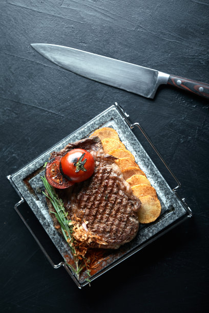 用刀叉雕刻在黑色石板上的烤牛排。在滚烫的大理石上烤牛排。复制空间，深色背景，美食时尚照片图片下载
