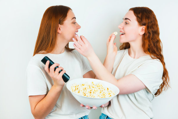 两个红发幸福的女人一个拿着遥控器喂对方爆米花站在孤立的白色背景，休闲时间的概念图片下载