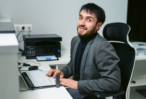 年轻的商人在办公室工作，坐在办公桌前，看着笔记本电脑屏幕，微笑着。图片下载