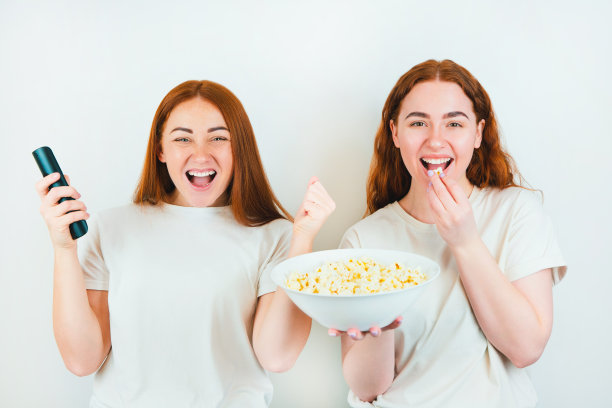 两个红发快乐的女人一个用遥控器另一个用爆米花看兴奋的电影在孤立的白色背景，休闲时间的概念图片下载