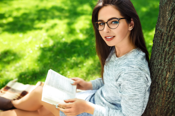 戴眼镜的漂亮女孩在夏天的公园里看书。图片下载