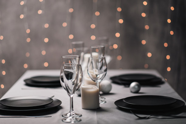 餐厅里摆好了一张桌子。一个表。灰色的艺术背景。黑色哑光器皿，玻璃玻璃。白色蜡烛和装饰。节日的黄色灯串在背景上。图片下载
