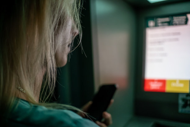 一名金发女孩手持手机站在ATM机附近，试图从卡中取钱，这是一张特写照片图片下载