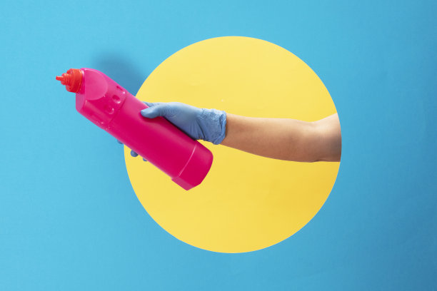 以蓝色和黄色纸为背景，手持装有洗涤剂瓶子的蓝色医用手套。图片下载