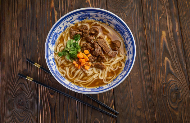中国牛肉面在传统的蓝色碗，新鲜的香草和胡萝卜片，一双筷子在深色的木制背景图片下载