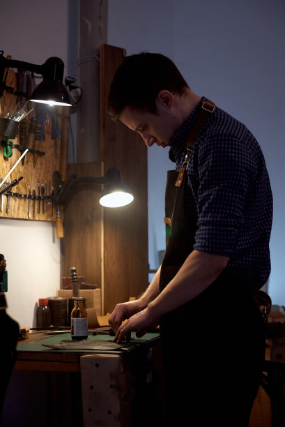 垂直照片的皮革工匠穿着一件蓝色的衬衫，站在桌旁和真正的棕色皮革工作。图片下载