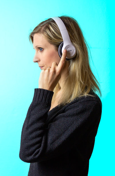美丽的年轻白种人金发女人的肖像听音乐耳机上的蓝色背景。女人按耳机上的按钮图片下载