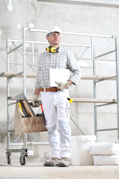 带着电脑和工具箱的建筑工人戴着手套、安全帽、眼镜和听力保护耳机，在有脚手架的建筑工地内部工作图片下载