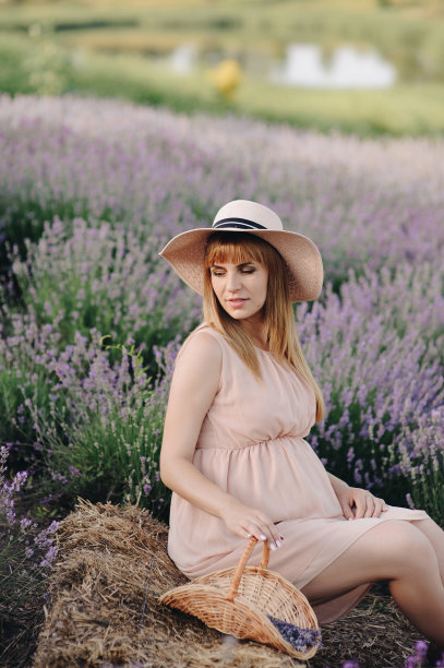 怀孕的金发女孩，米黄色的衣服和草帽。薰衣草。期待孩子的到来。拍摄照片的想法。走在日落时分。未来的妈妈。篮子里的花。图片下载