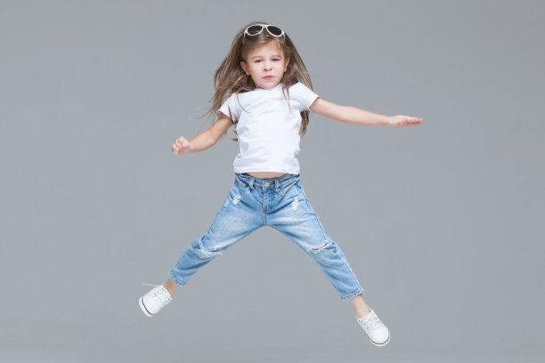 学龄前的小女孩穿着蓝色牛仔裤，白色t恤和太阳镜，举起的手孤立在灰色背景上跳跃图片下载