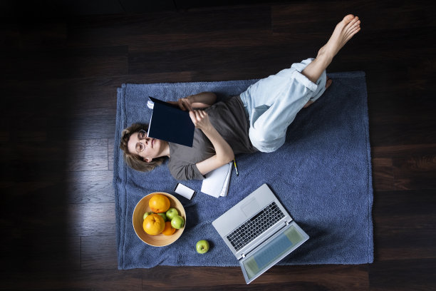年轻的女性博主或自由职业者放松躺在地板上的格子阅读旁边的笔记本电脑和一个水果篮顶视图图片下载