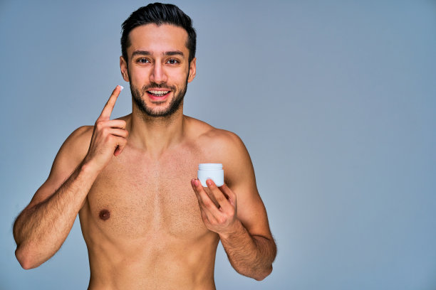 一个人微笑着，手里拿着一瓶面霜，在脸颊上涂着面霜。化妆品的概念图片下载