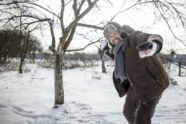 快乐的，微笑的男人，穿着敞开的夹克，围巾和帽子，玩雪球，正要扔一个雪球。冬天的乐趣。图片下载