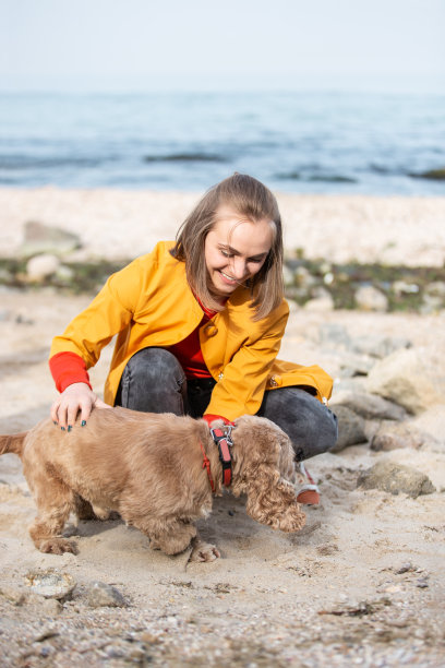 穿着黄色大衣的年轻女子在海边牵着狗散步。图片下载