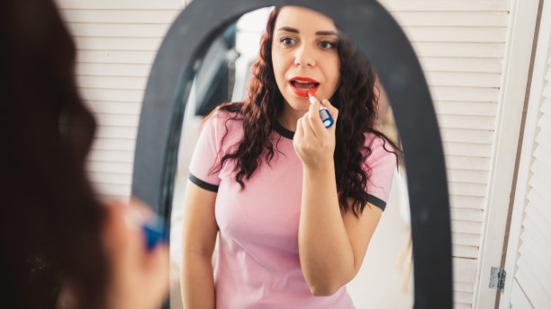 年轻漂亮的女人穿着睡衣涂上红色的口红，在家照镜子。迷人的女性在大镜子前梳妆的特写图片下载