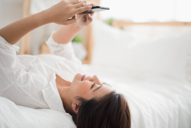 年轻的亚洲女人躺在家里白色的床上聊天和使用智能手机感觉快乐。人们在家里工作图片下载