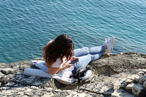 红头发的年轻女子在美丽的蓝色大海附近的石头海滩上晒太阳，看着智能手机图片下载