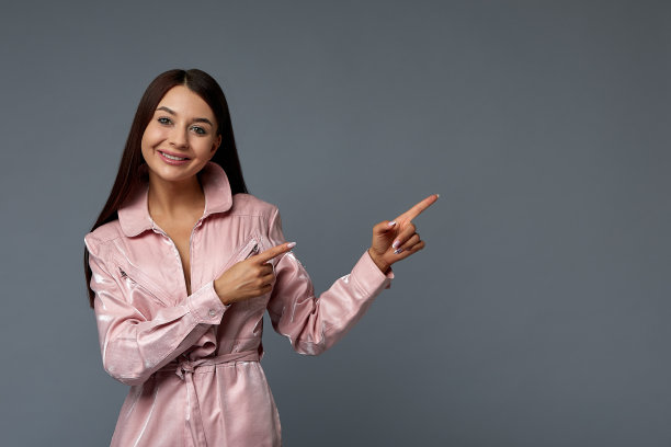 照片肖像，一个可爱的女人女孩与深色头发在粉红色的衣服在一个灰色的背景在工作室手指向复制空间。她一边说，一边微笑着把手放在镜头前。拷贝空间，灰色背景图片下载