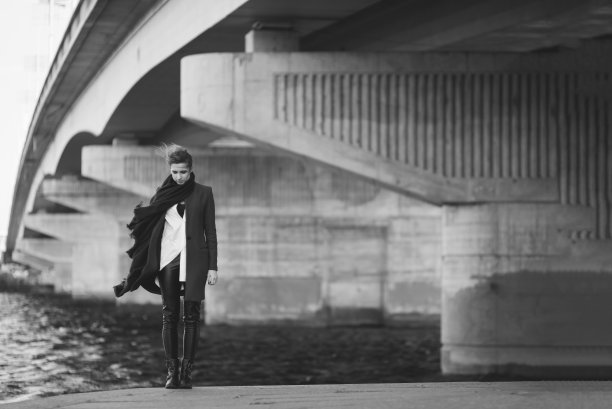 穿着大衣和围巾的美丽孤独的女人在河边的桥下随风飘动。孤独的概念。黑白艺术照片。软的焦点。图片下载