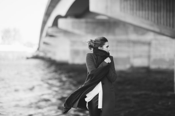 穿着大衣和围巾的美丽孤独的女人在河边的桥下随风飘动。孤独的概念。黑白艺术照片。软的焦点。图片下载
