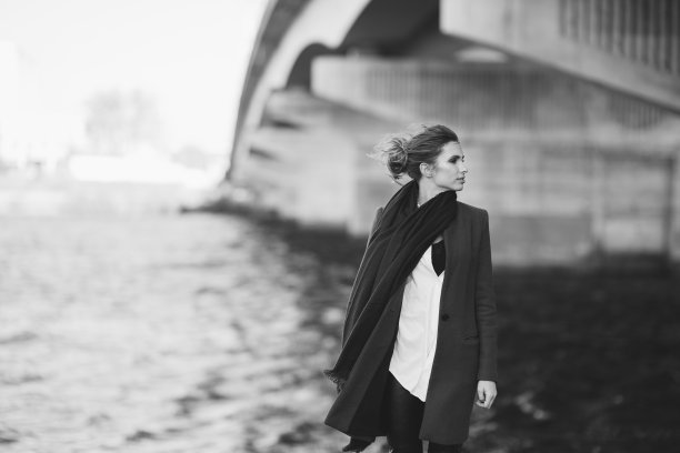 穿着大衣和围巾的美丽孤独的女人在河边的桥下随风飘动。孤独的概念。黑白艺术照片。软的焦点。图片下载