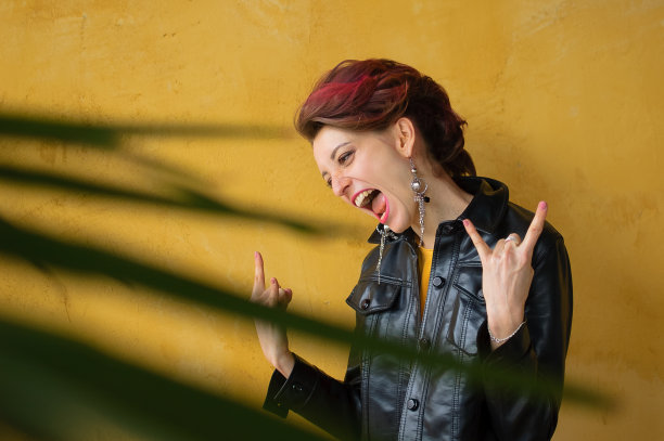 让我们摇滚的概念。兴奋的美丽女人的肖像与深粉红色的头发在休闲的黄色连衣裙站着，看着相机，显示摇滚手势。图片下载