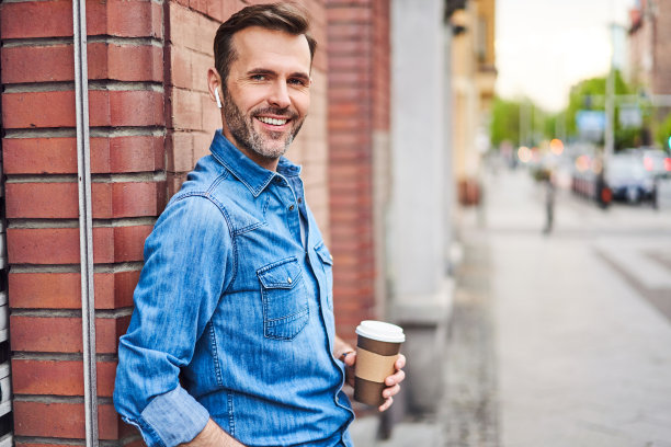 一名男子靠在墙上，戴着无线耳机边喝咖啡边打电话图片下载