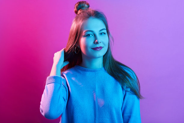 少女站立并触摸她美丽的秀发照片，扎着发髻，穿着休闲，在粉红色霓虹灯背景下摆姿势，看着镜头，看起来很自信。人物概念。图片下载