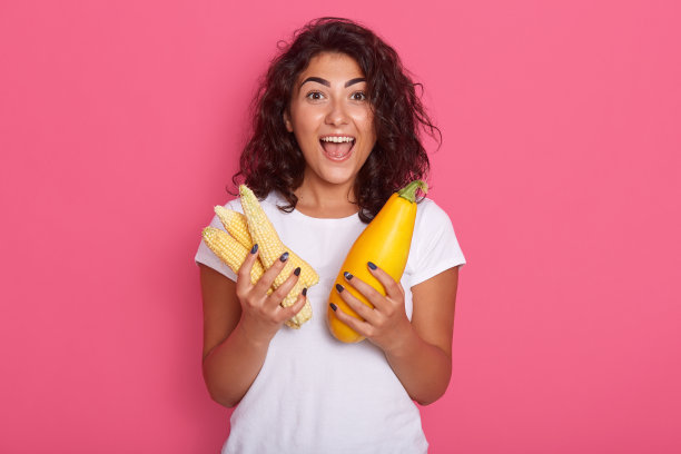 水平图片情绪良好的深色头发，张开的嘴和眼睛广泛，双手捧着西葫芦和玉米，穿着t恤，孤立地站在工作室的粉红色背景。图片下载
