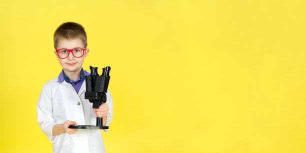 男孩戴着眼镜，穿着病号服，黄色背景上有显微镜。小科学家做发现，做实验。寻找治愈方法，病毒疫苗。未来的概念。横幅图片下载