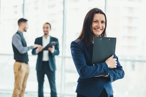 漂亮的办公室女员工的肖像，手里拿着文件夹，对着镜头可爱地微笑，她的同事们在模糊的背景上讨论着什么。图片下载