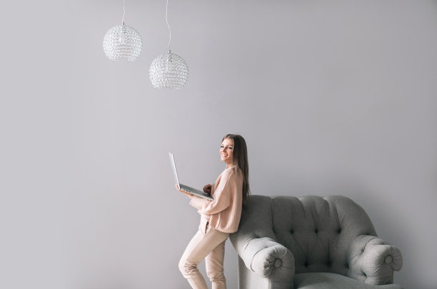 一位年轻漂亮的女士站在客厅的椅子旁边，用笔记本电脑工作。自由职业者。图片下载