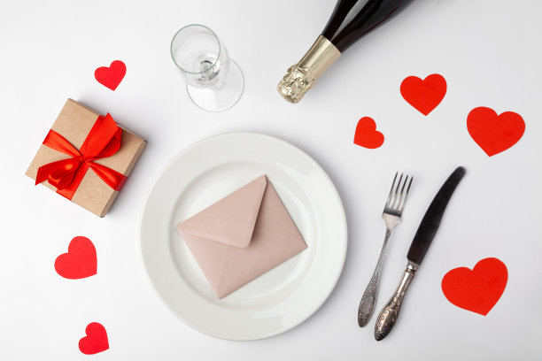 情人节晚餐与节日餐桌设置，红胶带，牛皮纸礼盒，香槟玻璃，香槟瓶，纸心，信封在盘子，银器在白色背景。前视图。图片下载