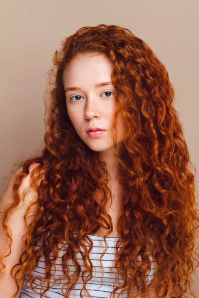 一个漂亮的模特看着镜头，她有着长长的红色卷发，棕色的眼睛和没有化妆的雀斑。工作室拍摄，皮肤修饰，中等拍摄，在米色背景。自然景观的概念图片下载